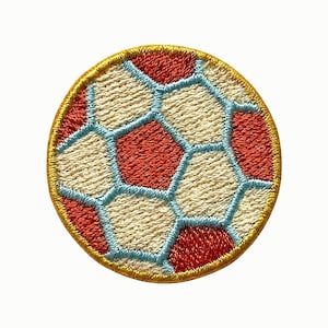 May include: A circular embroidered patch featuring a soccer ball design. The pattern consists of alternating red and off-white hexagonal and pentagonal shapes outlined in light blue thread, all enclosed by a solid gold-colored stitched border.