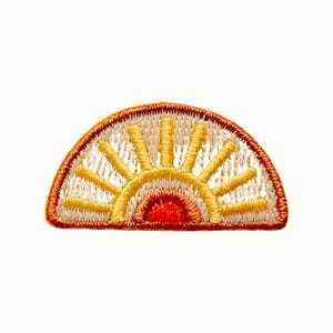 May include: A semi-circular embroidered patch featuring a stylized sun design. The center is a solid red semicircle, surrounded by concentric layers of yellow and cream-colored rays, all enclosed within a thick, dark orange stitched border.