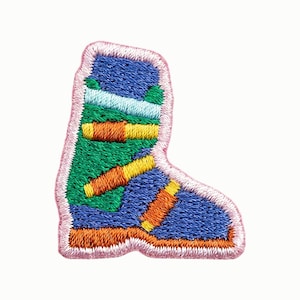 May include: An embroidered patch shaped like a ski boot with a light pink border. The boot features a blue upper section, a green mid-section, and a blue lower section with an orange sole. Three orange buckles are visible across the boot.