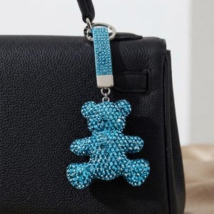 Puede incluir: Un colgante para bolso con forma de oso azul brillante cubierto de cristales centelleantes, unido a una correa texturizada azul a juego con un anillo de metal plateado. El colgante cuelga del asa de un bolso de cuero negro con textura.