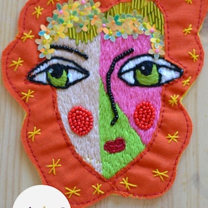 May include: An embroidered fabric patch featuring a stylized face with split-colored skin in beige, pink, and lime green. The face has green eyes, red beaded cheeks, and a red mouth. It is decorated with yellow star stitches and floral sequins.