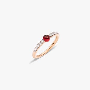 Könnte beinhalten: Ein Ring aus Roségold mit einem zentralen, rund geschliffenen roten Granat-Cabochon, der von einer Reihe kleiner, klarer, rund geschliffener weißer Diamanten flankiert wird, die beidseitig des Mittelsteins in die Ringschiene eingefasst sind.