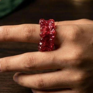 Könnte beinhalten: Ein durchscheinender, tiefroter Ring mit einem geschnitzten Blumenmuster auf der gesamten Oberfläche. Das Band hat eine dicke, strukturierte Form mit sich wiederholenden Blumenmotiven, die ein durchgehendes, skulpturales Design bilden.
