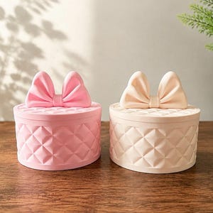 May include: Two round storage containers with lids, one in light pink and one in cream. Each lid features a prominent decorative bow. The sides of both containers have a raised, quilted diamond pattern texture across the entire exterior surface.