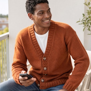 Op de afbeelding: Een gebreid vest in gebrande oranje kleur met een V-hals, lange mouwen met geribbelde boorden en een knoopsluiting aan de voorzijde met vier zichtbare bruine knopen. Het kledingstuk wordt gedragen over een wit T-shirt en een blauwe spijkerbroek.
