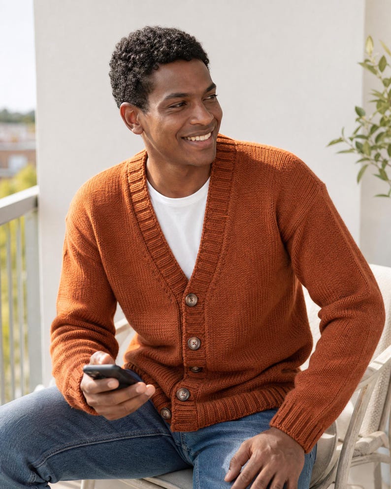 Op de afbeelding: Een gebreid vest in gebrande oranje kleur met een V-hals, lange mouwen met geribbelde boorden en een knoopsluiting aan de voorzijde met vier zichtbare bruine knopen. Het kledingstuk wordt gedragen over een wit T-shirt en een blauwe spijkerbroek.