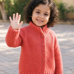 May include: A child wearing a long-sleeved, coral-colored knit cardigan with a ribbed mock neck collar, ribbed cuffs, and a ribbed hem. The cardigan features a vertical front placket with four matching coral-colored buttons and a textured knit pattern.