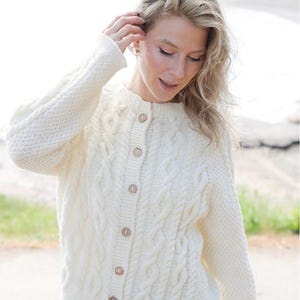 May include: A cream-colored, long-sleeved knit cardigan featuring a vertical cable knit pattern on the front panels and a textured stitch on the sleeves. The garment has a round neckline and is fastened down the center with five light brown buttons.