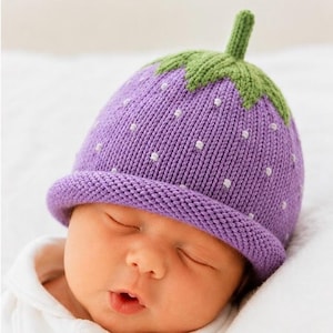 May include: A purple knit hat featuring a green stem at the top and small white embroidered dots across the surface. The hat has a rolled brim and a textured knit construction, designed to resemble a berry with a stem and seeds.
