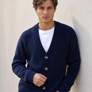 Op de afbeelding: Een marineblauw gebreid vest met lange mouwen, een V-hals, een knoopsluiting aan de voorzijde met vier zichtbare knopen en geribbelde details bij de kraag, manchetten en zoom, gedragen over een effen wit T-shirt met ronde hals.