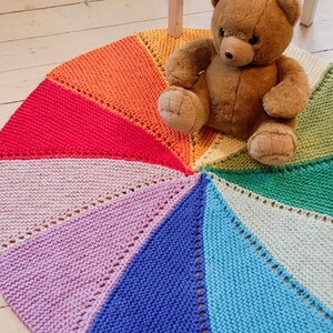 May include: A circular, hand-knitted rug featuring a vibrant rainbow color palette arranged in wedge-shaped segments. The rug displays a textured garter stitch pattern with eyelet details along the seams, accompanied by a brown plush teddy bear.