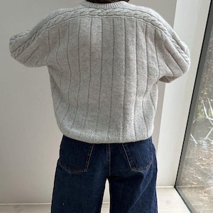 May include: A light gray knit sweater featuring a vertical ribbed pattern and cable knit detailing along the shoulders and sleeves, paired with dark blue denim jeans. The sweater has a crew neckline and long sleeves with a textured finish.