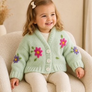 May include: A light green knit cardigan featuring a shawl collar, three white buttons, and embroidered floral motifs in pink, purple, and green. The long-sleeved sweater has ribbed cuffs and a ribbed hem, showcasing a soft, textured yarn construction.