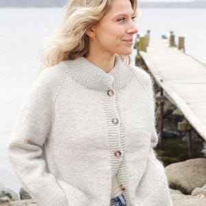 May include: A light grey, long-sleeved, knitted cardigan featuring a ribbed collar, cuffs, and hem. The garment is fastened with five brown, marbled buttons down the front placket. The knit texture appears soft and slightly fuzzy in appearance.