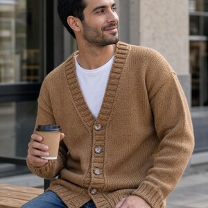 Può includere: Un cardigan in maglia color marrone chiaro con scollo a V, maniche lunghe e chiusura frontale con quattro bottoni marroni. Il capo è indossato sopra una t-shirt bianca semplice e abbinato a jeans blu, mentre si tiene in mano un bicchiere di caffè usa e getta.