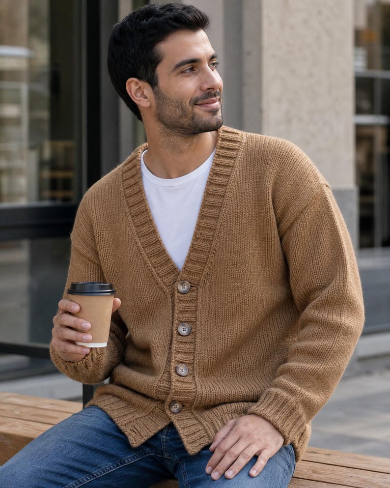 Op de afbeelding: Een beige gebreid vest met een V-hals, lange mouwen en een knoopsluiting aan de voorzijde met vier bruine knopen. Het kledingstuk wordt gedragen over een effen wit T-shirt en gecombineerd met een blauwe spijkerbroek, terwijl een wegwerpbeker wordt vastgehouden.