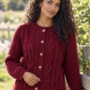 May include: A deep red cable-knit cardigan featuring a round neckline, long sleeves with ribbed cuffs, and a front button closure with five light-colored circular buttons. The garment displays a textured pattern of vertical cables and side panels.