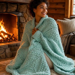 May include: A light mint green knitted throw blanket featuring a textured pattern of raised bobbles and diamond-shaped eyelet details. The blanket is crafted with a thick yarn, showcasing a complex geometric design across its entire surface area.