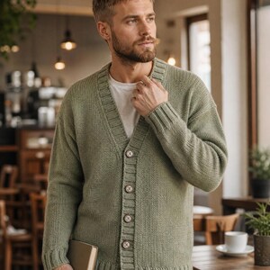 Op de afbeelding: Een saliegroen gebreid vest met een V-hals, lange mouwen met geribbelde boorden en een knoopsluiting aan de voorzijde met vier zichtbare bruine knopen. Het kledingstuk wordt gedragen over een effen wit T-shirt en gecombineerd met een blauwe spijkerbroek.