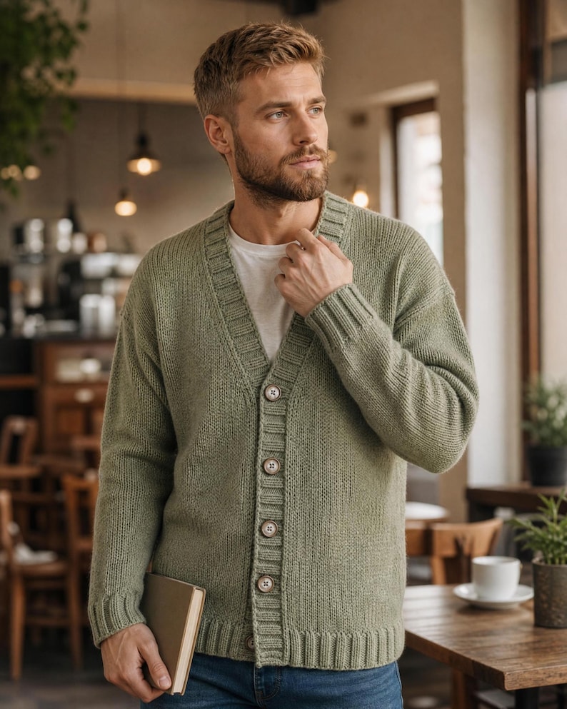 Op de afbeelding: Een saliegroen gebreid vest met een V-hals, lange mouwen met geribbelde boorden en een knoopsluiting aan de voorzijde met vier zichtbare bruine knopen. Het kledingstuk wordt gedragen over een effen wit T-shirt en gecombineerd met een blauwe spijkerbroek.