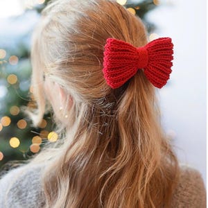 Op de afbeelding: Een felrode gebreide haarstrik met een geribbelde textuur en een centraal knoopdetail. Het accessoire is ontworpen om in het haar te dragen en toont een klassieke strikvorm met duidelijke horizontale gebreide lijnen over het oppervlak.