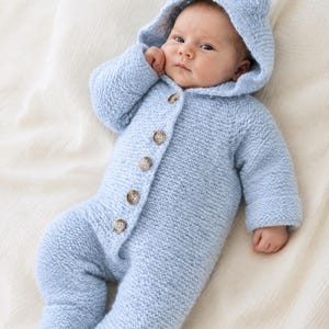 May include: A light blue knitted baby romper featuring a hood with two small rounded ears on top. The garment has long sleeves, full-length legs, and a front closure secured by five brown buttons. The texture of the knit is consistent throughout.