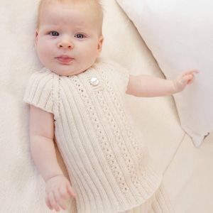 Könnte beinhalten: Ein cremefarbenes, gestricktes Baby-Oberteil und eine passende Shorts mit vertikalem Rippenmuster und zarten Lochmuster-Details. Das Oberteil hat kurze Ärmel, einen Rundhalsausschnitt und einen einzelnen weißen Knopf in der Brustmitte.