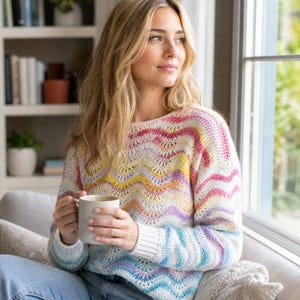 May include: A long-sleeved crochet sweater featuring a wavy, multi-colored pattern in shades of pink, yellow, orange, purple, and blue on a cream-colored base. The sweater has a round neckline and ribbed cuffs at the wrists, with a textured stitch.