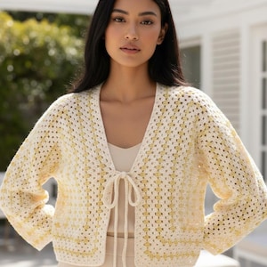 May include: A cropped crochet cardigan featuring a granny square pattern in cream and pale yellow. The garment has long sleeves, an open front design, and a thin matching tie closure at the center of the neckline. The knit texture is visible throughout.