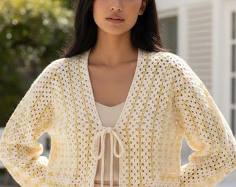 Op de afbeelding: Een kort gehaakt vest met een granny square patroon in crème en lichtgeel. Het kledingstuk heeft lange mouwen, een open voorzijde en een dunne bijpassende striksluiting in het midden van de halslijn. De gebreide textuur is overal zichtbaar.