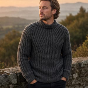 May include: A dark charcoal grey long-sleeve jumper featuring a thick ribbed knit texture, a high mock neck collar, and raglan sleeves. The garment is paired with dark-coloured trousers, showcasing a heavy knit construction and a fitted silhouette.