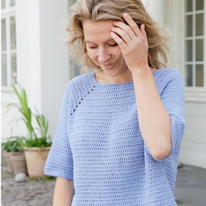 May include: A light blue crochet top featuring a round neckline and short sleeves. The garment is constructed with a consistent horizontal stitch pattern throughout, including a subtle decorative diagonal line of openwork stitches near the shoulder.