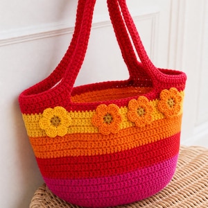 Op de afbeelding: Een gehaakte tas met horizontale strepen in de kleuren rood, oranje, geel en roze. De tas heeft twee rode hengsels en is aan de bovenzijde aan de voorzijde versierd met vier gehaakte bloemapplicaties in geel en oranje.