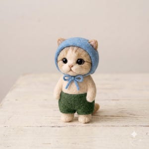 Puede incluir: Una figura de gatito de lana fieltrada con pelaje beige claro y grandes ojos negros. El gatito lleva un gorro azul suave atado con un lazo bajo la barbilla y pantalones cortos de color verde oscuro. La figura tiene cabeza redonda y cuerpo compacto.