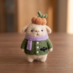 May include: A needle-felted beige animal figurine wearing a green cardigan with three white buttons and a black bat embroidery. The figure has a lavender scarf around its neck and a small orange pumpkin with a green stem resting on its head.