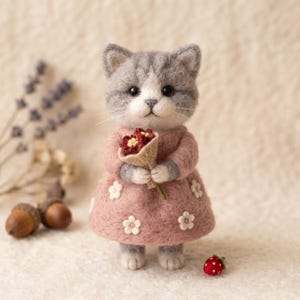May include: A needle-felted gray and white kitten figurine wearing a dusty pink dress adorned with small white flower appliques. The kitten holds a tiny bouquet of dark red flowers wrapped in beige fabric, with a small red strawberry nearby.