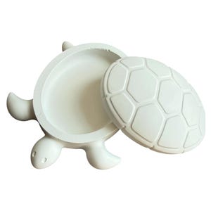 May include: A white turtle-shaped trinket box with a removable lid. The base features a hollow interior and the lid is molded with a raised hexagonal shell pattern. The object is crafted from a solid white material with a smooth, matte finish.