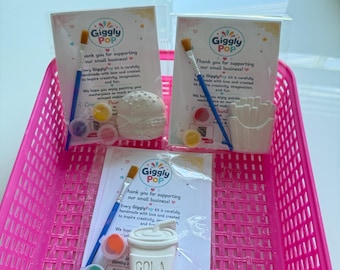 May include: Three GigglyPop craft kits in clear plastic packaging, each containing a white plaster figure of a burger, fries, or a cola cup, a blue paintbrush, and small paint pots, all displayed inside a bright pink plastic storage basket.