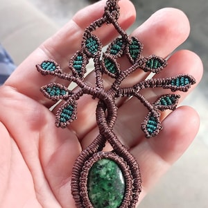 Könnte beinhalten: Ein Makramee-Anhänger mit einem Baumdesign aus brauner Kordel und kleinen blaugrauen Blattakzenten. Die Basis des Baumes enthält einen glatten, ovalen grünen Stein mit dunklen Sprenkeln, eingefasst in einen Rahmen aus brauner Kordel.