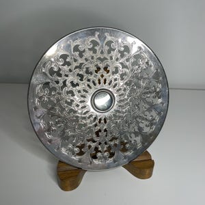 May include: A circular silver-toned metal plate featuring an intricate, openwork filigree pattern of scrolling vines and floral motifs radiating from a smooth, solid centre disc, displayed upright on a small, brown wooden stand.
