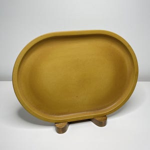 Può includere: Un vassoio ovale in ceramica color giallo senape con una finitura liscia e opaca e un bordo rialzato. Il vassoio è esposto in verticale, sostenuto da un piccolo supporto in legno a due punte che mantiene la base in posizione verticale.