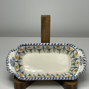 May include: A rectangular ceramic serving dish with rounded corners and integrated handles. The interior features a cream-colored base decorated with a repeating pattern of blue and gold swirls and lines along the inner rim and handle edges.