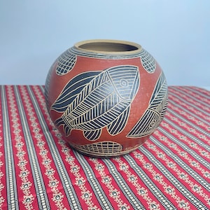 May include: A round, terracotta-coloured ceramic vase featuring hand-etched black fish motifs with intricate line detailing. The vessel has a wide, open mouth and a smooth, matte finish, with the artist's signature visible near the base.