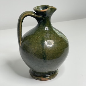 May include: A small, rounded ceramic pitcher with a glossy, deep forest green glaze. The vessel features a single curved handle on the side, a narrow neck, and a small pouring spout. The base is circular and slightly flared, showing hints of brown clay.