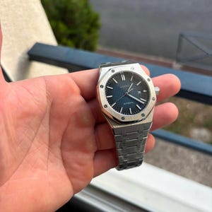 May include: A silver-toned stainless steel wristwatch featuring an octagonal bezel with eight visible screws, a dark blue textured dial with a grid pattern, silver hour markers, silver hands, and a matching integrated link bracelet strap.
