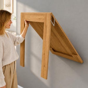 May include: A light oak wood wall-mounted folding table featuring a rectangular surface and two vertical legs. The table is shown in a partially folded position against a gray wall, with a metal hinge mechanism visible on the side of the frame.