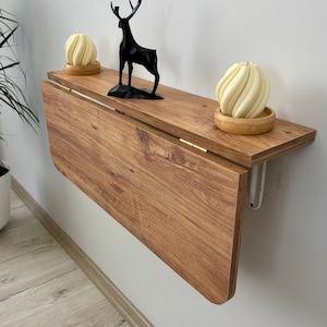 May include: A wall-mounted wooden drop-leaf table with a light brown wood grain finish. The table features a hinged top section that folds downward. On the surface sit two cream-coloured spiral candles in wooden bases and a black deer figurine.