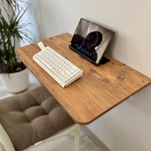 May include: A wall-mounted wooden desk with a light brown wood grain finish. On the desk surface sits a white mechanical keyboard and a tablet computer in a black case. A brown tufted seat cushion is positioned on a chair beneath the desk.