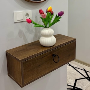 Puede incluir: Un estante flotante con cajón en acabado de madera oscura y un tirador de anillo circular negro. Sobre él, un jarrón de cerámica blanca con forma de doble bulbo acanalado contiene un ramo de tulipanes artificiales en tonos rojos, rosas, amarillos y morados.