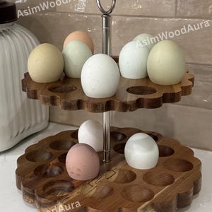 May include: A two-tiered wooden egg holder with a scalloped edge design and a central silver metal handle. The stand features multiple circular indentations for holding eggs, with several multi-colored eggs placed on both levels. Text reads AsimWoodAura.